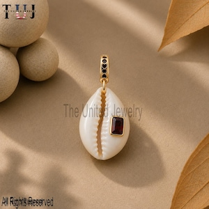 May include: A white cowrie shell pendant featuring a gold-toned metal bail and a rectangular, deep red gemstone set in a gold-toned frame on the front. The pendant is suspended from a gold-toned loop with a series of small, dark circular accents.