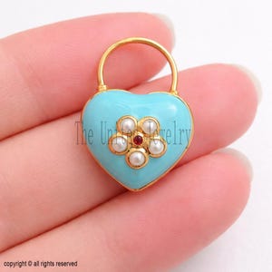 May include: A heart-shaped pendant with a light blue enamel finish and a gold-colored bail. The pendant features a flower design made of white pearls and a central red gemstone. The text "The United Jewelry" is visible on the pendant.