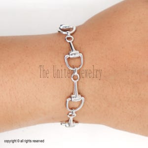 Può includere: Un braccialetto argentato con un design a morso di cavallo. Il braccialetto è composto da diversi anelli a forma di morso collegati. Il testo "The United Jewelry" è visibile sullo sfondo. Il braccialetto è indossato al polso.