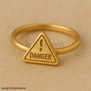 May include: A gold-colored ring featuring a triangular danger sign with an exclamation point and the word "DANGER" in capital letters. The ring has a smooth, polished finish and a simple band.