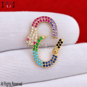 May include: A gold-toned carabiner-style jewellery clasp featuring a multi-coloured pavé design. The surface is encrusted with small, vibrant cubic zirconia stones in shades of pink, purple, white, green, turquoise, blue, and black, arranged in a gradient pattern along the curved frame.