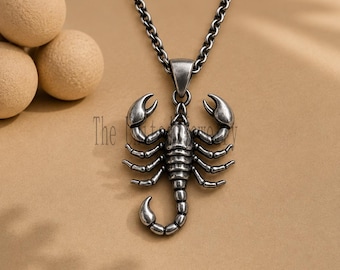 May include: A silver-toned metal scorpion pendant suspended from a matching link chain. The pendant features a detailed, textured scorpion design with raised pincers and a curved tail. The metal has an oxidized finish, highlighting the segments.