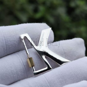 Pode incluir: Um pingente prateado em forma da letra "K" com um pequeno detalhe retangular dourado. O pingente é segurado em uma luva branca. O texto "The United Jewelry" é visível.