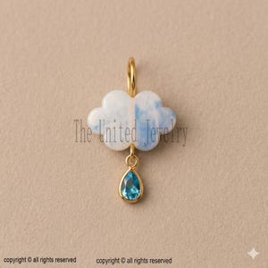 Puede incluir: Un colgante dorado con forma de nube con una lágrima azul. La nube es blanca y azul claro. El colgante tiene un lazo en la parte superior para una cadena. La lágrima es una piedra preciosa azul claro.