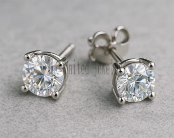 May include: A pair of silver stud earrings with round, clear gemstones. Each earring has a four-prong setting holding a sparkling, brilliant-cut stone. The earrings are displayed on a light grey surface.