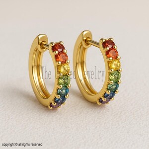 Puede incluir: Un par de pendientes de aro dorados con un arcoíris de piedras preciosas. Las piedras preciosas están dispuestas en una fila de rojo, naranja, amarillo, verde, azul y morado. Un accesorio colorido y clásico.