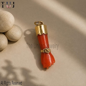 Puede incluir: Un colgante con forma de puño tallado en coral de color rojo anaranjado, que presenta una tapa de metal dorado en la parte superior con una anilla y un anillo de metal dorado que rodea la zona de la muñeca junto a tres piedras lisas.