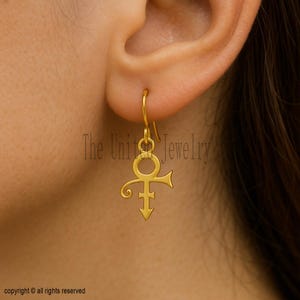 May include: A gold-toned dangle earring featuring the Prince love symbol, which consists of a circle with a cross and an arrow pointing downward. The earring hangs from a simple hook wire and is displayed against an earlobe for scale.