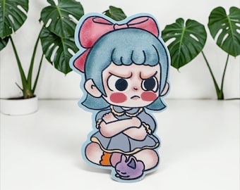 May include: A sticker depicting a cartoon girl with blue hair and a pink bow, with a stern expression and crossed arms. She wears a grey dress and has a purple cat at her feet. The sticker has a light blue border.