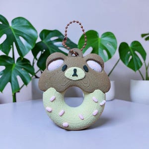 Puede incluir: Un llavero con forma de dónut de oso en color marrón y verde claro con virutas rosas y una superficie texturizada. El oso tiene un hocico color crema, nariz negra y el interior de las orejas rosa. Una cadena de bolas marrón está unida.