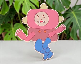 May include: A cartoon sticker of a character with a pink head and body, pink arms, and blue legs. The character has a round face with an angry expression. The sticker is set against a backdrop of green leaves.