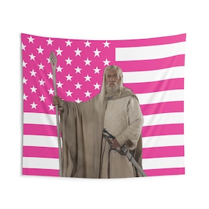 May include: A tapestry featuring a character from a fantasy film, holding a staff and sword, set against a pink and white American flag design. The character is dressed in beige robes.