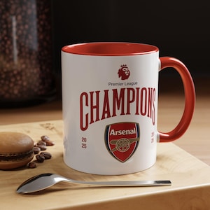 Könnte beinhalten: Weißer Keramikbecher mit rotem Innenraum und Henkel. Der Becher zeigt den Text "CHAMPIONS" in Rot, zusammen mit dem Arsenal-Logo und dem Text "Premier League". Ein Löffel und ein Macaron liegen auf einer Holzoberfläche.