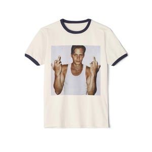 May include: Cream-colored ringer t-shirt with a navy blue collar and sleeve trim. The shirt features a photographic print of a person wearing a white tank top, raising both middle fingers.