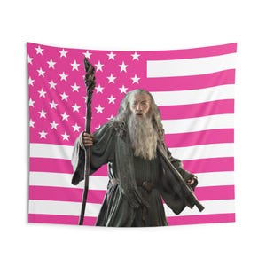 May include: A tapestry featuring a pink and white American flag design with a portrait of a wizard holding a staff. The wizard has a long white beard and is wearing a gray robe and scarf.