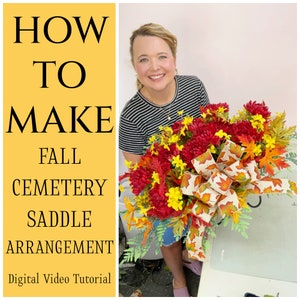May include: How to Make Fall Cemetery Saddle Arrangement - Digital Video Tutorial. A woman holds a fall floral arrangement with red, yellow, and orange flowers, a bow, and greenery.