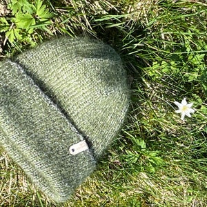 May include: A green knitted beanie hat with a brown leather label. The hat is lying on a bed of green grass.