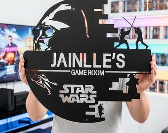 Könnte beinhalten: Ein schwarzes Metallschild im Star Wars-Design mit dem Text "JAINLLE'S GAME ROOM". Das Schild zeigt Silhouetten von Charakteren und Elementen aus dem Star Wars-Franchise, darunter Darth Vader und Lichtschwertduelle. Das Schild wird von einer Person gehalten.
