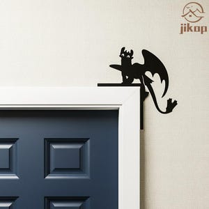 May include: Black metal dragon silhouette decorates a door frame. The dragon has large wings and a long tail. The door is navy blue with a white frame. The wall is a light beige color. The dragon is a decorative accent.