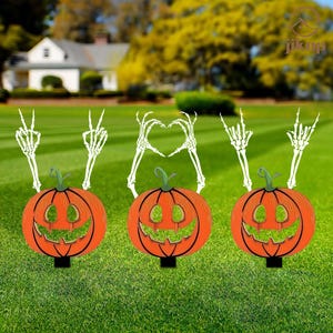 Può includere: Tre decorazioni da giardino a forma di zucca arancione con facce sorridenti verdi e basi nere. Mani di scheletro bianche sono posizionate sopra ogni zucca, formando segni di pace e una forma di cuore. Le zucche sono su un prato verde.
