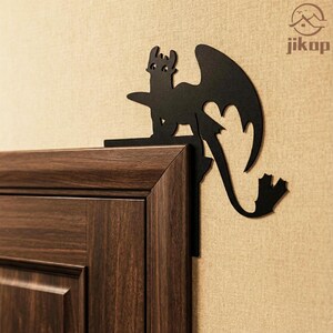 May include: A black metal dragon silhouette decorates a wooden door frame. The dragon has large wings and a long tail. The dragon is perched on the corner of a dark brown door frame against a beige wall. The logo "jikop" is in the upper right corner.