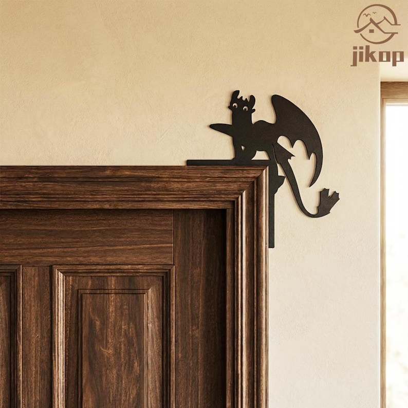 May include: A black metal dragon silhouette decorates a door frame corner. The dragon has large wings and a long tail. The door is made of dark brown wood. The wall is a light beige color. The dragon is a decorative accent.