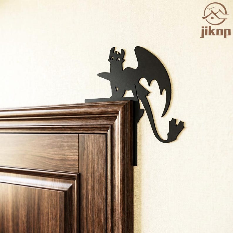 May include: Black metal dragon silhouette decor, designed to sit on a corner. The dragon has large wings and a long tail. The decor is mounted on a dark brown wooden door frame and a cream-colored wall.