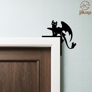 May include: Black dragon silhouette decor, perched on a door frame. The dragon has large wings and a long tail. The decor is mounted on a white door frame and a brown door. The wall is light blue. The logo "jikop" is in the upper right corner.