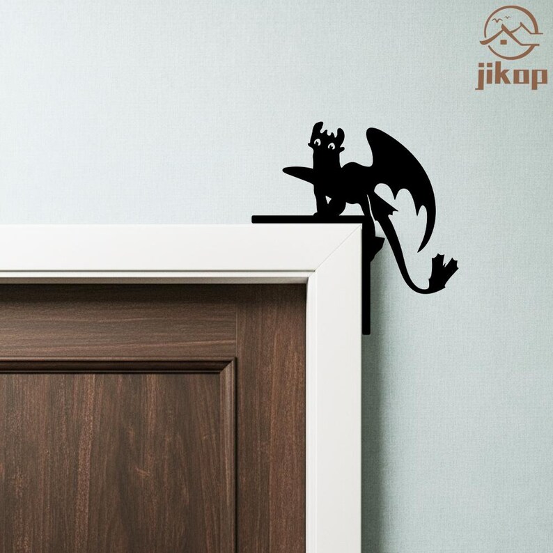 May include: Black dragon silhouette decor, perched on a door frame. The dragon has large wings and a long tail. The decor is mounted on a white door frame and a brown door. The wall is light blue. The logo "jikop" is in the upper right corner.