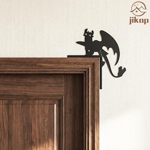 May include: Black metal dragon corner decor. The dragon is a silhouette with outstretched wings and a long tail. It is mounted on the corner of a wooden door frame. The dragon is a decorative accent piece.