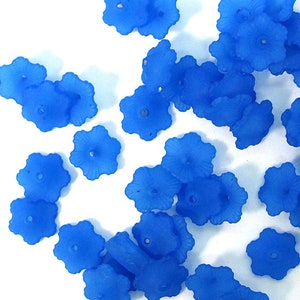 May include: A collection of small, blue, flower-shaped beads scattered on a white surface. Each bead has a six-petal design with a small hole in the center, suggesting they are for stringing or crafting. The beads are a vibrant, solid blue color.