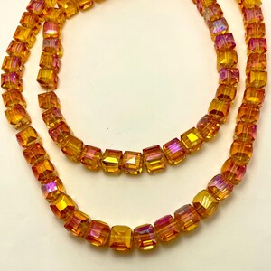 May include: Two strands of iridescent, cube-shaped beads in shades of yellow, orange, and pink. The beads are strung together, creating a necklace-like appearance. The beads have a faceted surface that catches the light.