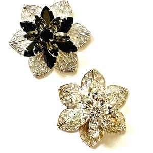 May include: Two flower-shaped brooches. One brooch features a silver-tone filigree design with black and gray crystals. The other brooch has a similar silver-tone filigree design with clear crystals. Both brooches are on a white background.