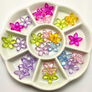 May include: A white ceramic divided tray filled with colorful, translucent flower-shaped beads. The beads are in shades of pink, purple, yellow, green, and blue. The flowers are arranged in separate compartments, showcasing their variety.