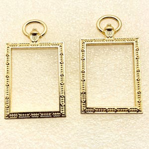 May include: Two gold-colored rectangular frames with decorative edges and a circular loop at the top. The frames have intricate detailing along the borders, with a repeating pattern of small shapes. The frames are empty in the center.