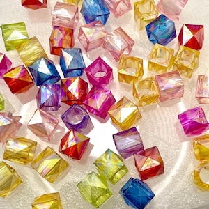 May include: An assortment of colorful, faceted beads in various shapes and sizes. The beads are in shades of yellow, green, blue, pink, and red, with an iridescent sheen. Some beads are cube-shaped, while others are ring-shaped. These beads are ideal for jewelry making.