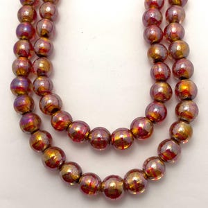 May include: Three strands of iridescent glass beads in shades of red, gold, and purple. The round beads have a glossy finish and are strung together, creating a necklace or jewelry component.