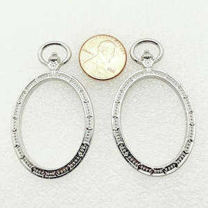 May include: Two silver-colored, oval-shaped jewelry findings with decorative edges and a loop at the top. A penny is placed between the findings for size comparison. The findings are on a white surface.