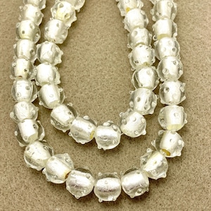 May include: Three strands of clear glass beads with a textured, bubbly surface. The beads have a silver-like interior and are strung together, creating a necklace-like appearance. The background is a neutral tan color.