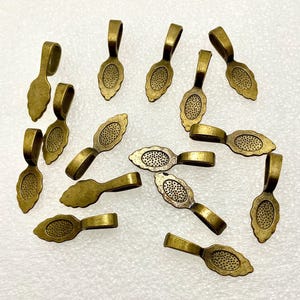 May include: A collection of antique brass-colored pendant bails. Each bail features a decorative oval with a dotted pattern and a loop for attaching to a chain or other jewelry component. The bails have a vintage, aged appearance.
