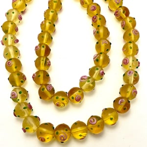 May include: A necklace featuring round, yellow glass beads. Each bead has pink and green decorative swirls. The necklace is displayed against a white background, highlighting the vibrant colors and intricate details of the beads.