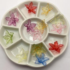 May include: A white ceramic divided tray filled with colorful, translucent flower-shaped embellishments. The flowers are in shades of red, pink, yellow, green, and blue. These decorative elements are likely for crafts or nail art.