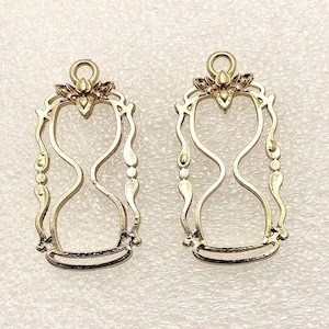 May include: Two gold-colored hourglass charms. Each charm features an hourglass shape within a decorative frame with floral accents at the top. The charms have a loop at the top for attaching to jewelry.