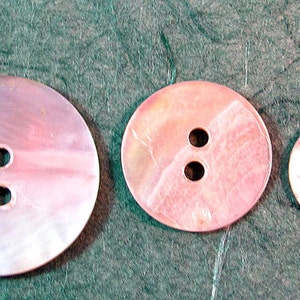 May include: Three iridescent pink mother-of-pearl buttons with two holes each. The buttons are oval shaped and have a smooth, shiny finish.