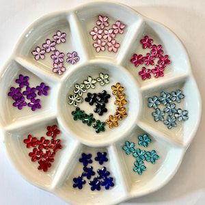 May include: A white ceramic tray filled with various colors of flower-shaped rhinestones. The rhinestones are in shades of pink, purple, red, blue, green, and gold. These decorative embellishments are ideal for crafts and nail art.