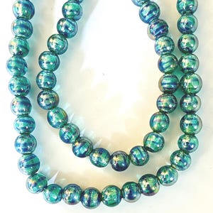 May include: A necklace featuring iridescent, round beads in shades of teal, green, and blue. The beads have a striped pattern and are strung together to form a multi-strand necklace. The necklace is displayed against a white background.