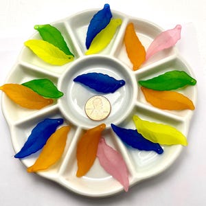 May include: A white ceramic divided tray filled with colorful, translucent leaf-shaped beads. The beads are in shades of blue, yellow, orange, green, and pink. A penny is in the center for size reference.
