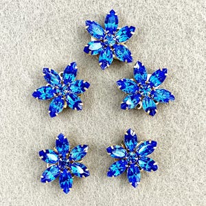 May include: Five blue rhinestone flower embellishments with gold-colored metal backing. Each flower has six petals and a central rhinestone. The rhinestones are various shades of blue, creating a sparkling effect. These are ideal for craft projects.