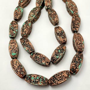 May include: A strand of cylindrical beads featuring a copper-toned finish with recessed black detailing and turquoise-colored accents. Each bead displays a repeating floral pattern across its surface, strung together on a thin white cord.