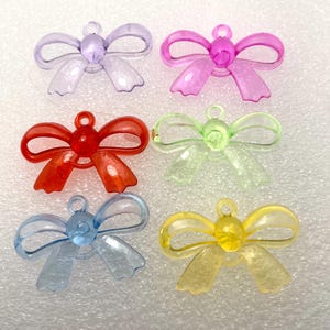 May include: Six translucent bow charms in various colors: purple, pink, red, green, blue, and yellow. Each bow has a loop for hanging. These charms are suitable for jewelry making or craft projects.
